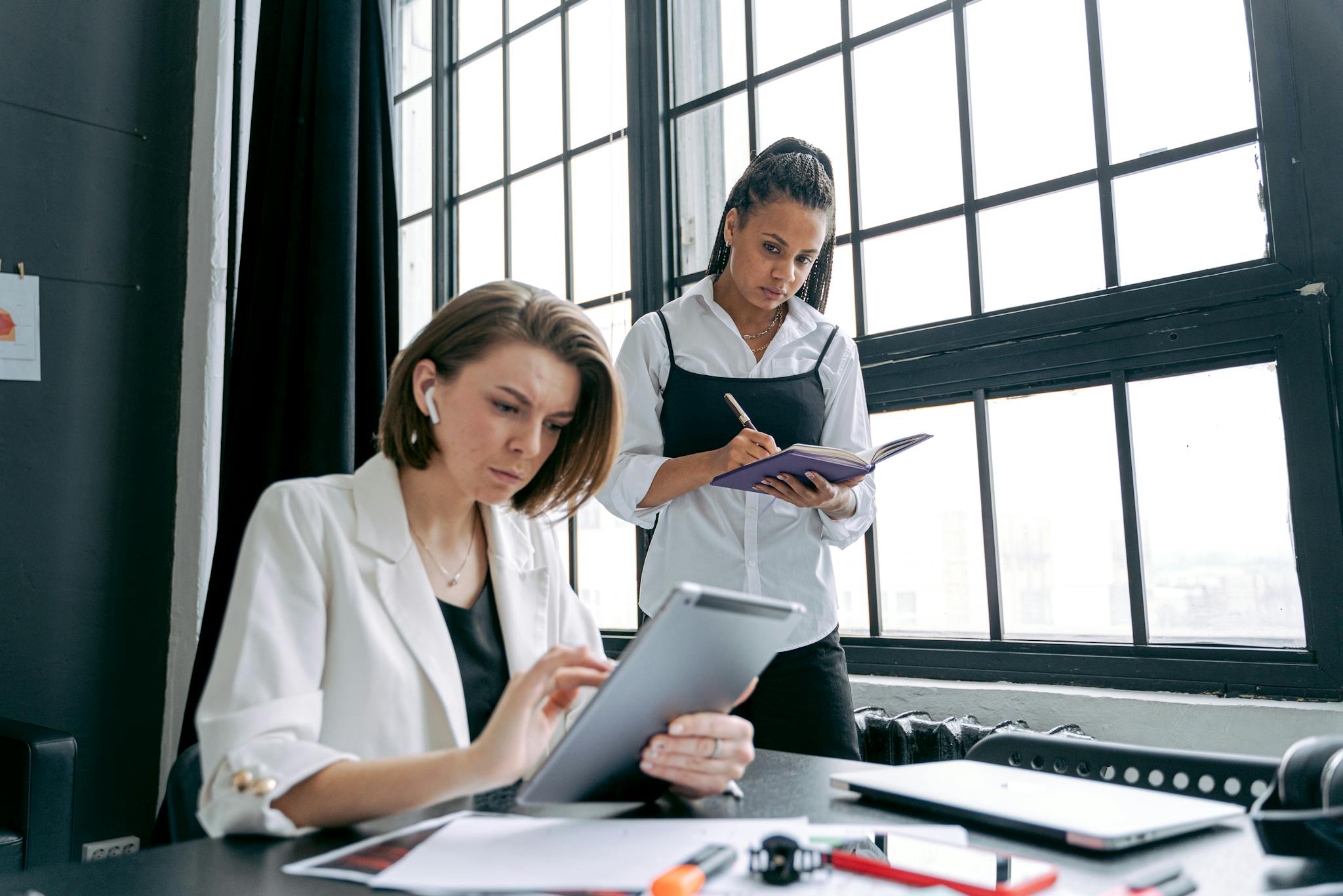 Two professional women working together in a modern office, using technology and documents.